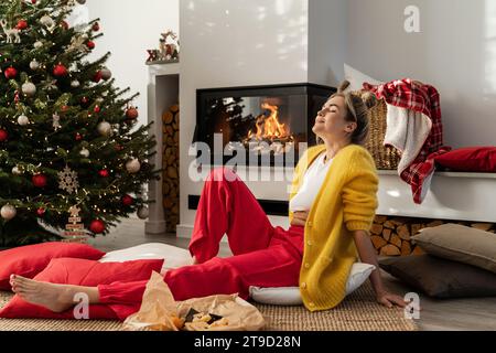 Jeune femme est assise à côté d'une cheminée lumineuse dans un salon confortable orné d'un sapin de Noël et de décorations festives. Banque D'Images