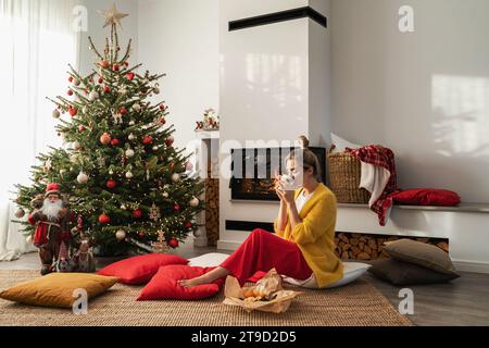 Femme heureuse assis à côté d'une cheminée incandescente dans un salon confortable, orné d'un arbre de Noël et de décorations festives, profitant d'une tasse de boisson chaude. Banque D'Images