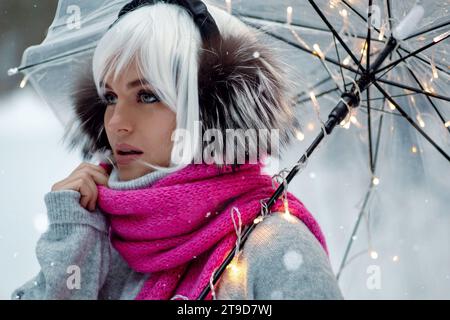 Femme, ornée d'une perruque blanche, cache-oreilles thermiques, pull confortable et écharpe rose. Elle tient un parapluie transparent encastré avec des lumières scintillantes, creati Banque D'Images