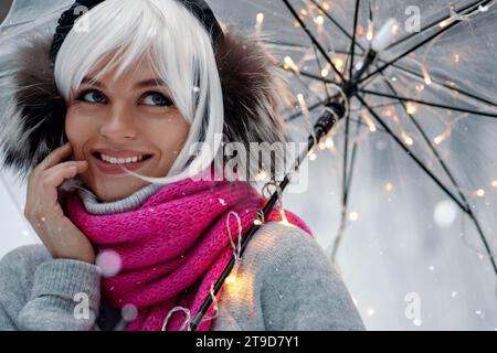 Femme, ornée d'une perruque blanche, cache-oreilles thermiques, pull confortable et écharpe rose. Elle tient un parapluie transparent encastré avec des lumières scintillantes, creati Banque D'Images