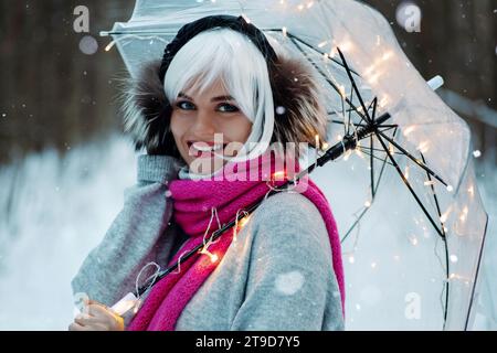 Femme, ornée d'une perruque blanche, cache-oreilles thermiques, pull confortable et écharpe rose. Elle tient un parapluie transparent encastré avec des lumières scintillantes, creati Banque D'Images