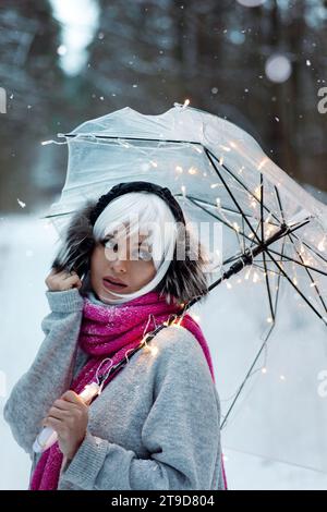 Femme, ornée d'une perruque blanche, cache-oreilles thermiques, pull confortable et écharpe rose. Elle tient un parapluie transparent encastré avec des lumières scintillantes, creati Banque D'Images