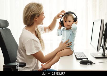 Jeune mère pigiste met ses écouteurs sans fil sur son petit fils assis au bureau de son bureau à domicile. Banque D'Images