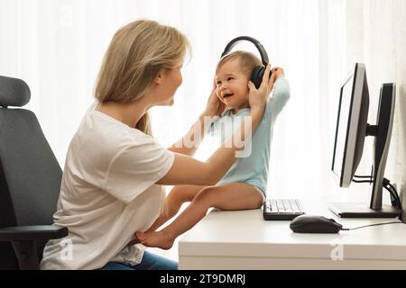 Jeune mère pigiste met ses écouteurs sans fil sur son petit fils riant assis au bureau de son bureau à domicile. Banque D'Images