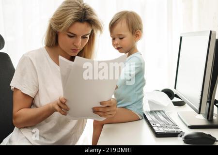 Jeune mère freelance lit quelques papiers à son lieu de travail de bureau à domicile avec son petit fils assis sur le bureau. Banque D'Images