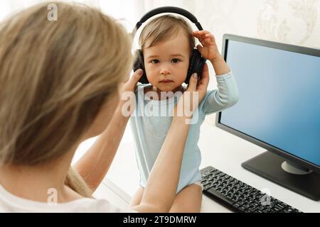 Jeune mère pigiste met ses écouteurs sans fil sur son petit fils assis au bureau de son bureau à domicile. Banque D'Images