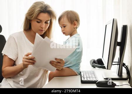 Jeune mère freelance lit quelques papiers à son lieu de travail de bureau à domicile avec son petit fils assis sur le bureau. Banque D'Images