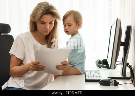 Jeune mère freelance lit quelques papiers à son lieu de travail de bureau à domicile avec son petit fils curieux assis sur le bureau. Banque D'Images