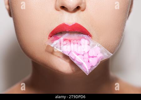 Plan en gros plan d'une jeune femme tenant un sac ziplock transparent rempli de pilules roses en forme de cœur dans sa bouche. Banque D'Images