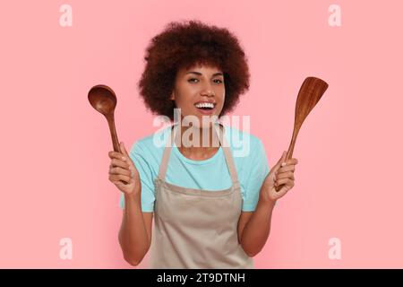 Heureuse jeune femme en tablier tenant cuillère et spatule sur fond rose Banque D'Images