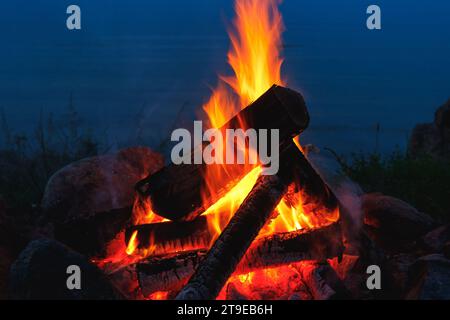Gros plan d'un feu de camp brûlant brillamment la nuit dans un camp en plein air avec une mer calme sur fond. Banque D'Images