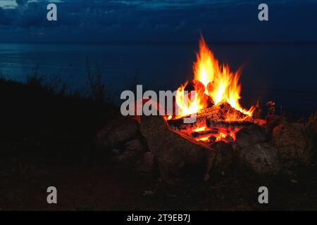 Gros plan d'un feu de camp brûlant brillamment la nuit dans un camp en plein air avec une mer calme sur fond. Banque D'Images