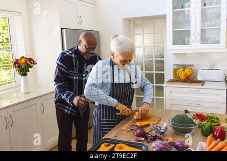 Heureux couple senior diversifié tranchant la courge butternut, hachant les légumes dans la cuisine Banque D'Images