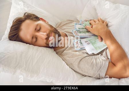 Homme endormi souriant couché dans son lit avec les yeux fermés, embrassant beaucoup de billets de banque en dollars à la maison Banque D'Images