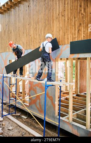 Charpentiers construisant une maison encadrée en bois. Deux hommes ouvriers claviant la façade de la maison avec des panneaux de particules de ciment, les fixant avec des tournevis. Concept de construction écologique moderne. Banque D'Images