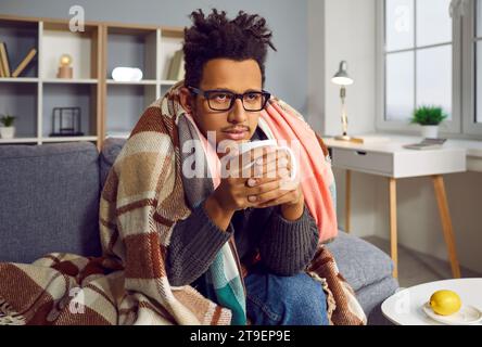 Triste homme afro-américain malade ayant un rhume, assis sur le canapé et buvant du thé chaud Banque D'Images