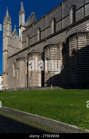 Côté sud de la cathédrale d'Orvieto, le Duomo di Santa Maria Assunta, en Ombrie, Italie. Les murs latéraux rayés de la cathédrale sont construits avec des couches de travertin blanc local et de pierre de basalte bleu-gris. Cette image montre également l'arrière de la façade ouest et ses tours avec des pinacles gothiques croqués. Banque D'Images