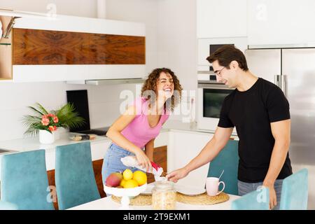 Couple positif s'amusant tout en se tenant près de la table à manger rire femelle avec les yeux fermés, verser du jus et plaisanter regardant vers le bas mâle dans des lunettes Banque D'Images