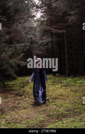 femme en robe noire et grise avec manches chauve-souris et crânes marchant vers la caméra dans une forêt Banque D'Images