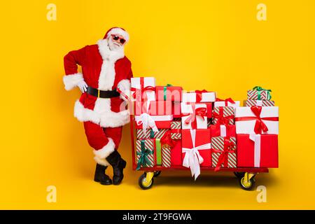 Photo pleine longueur de père noël vieilli positif pousser grand pile pile pile chariot de boîte-cadeau nouvelle année isolé sur fond jaune Banque D'Images