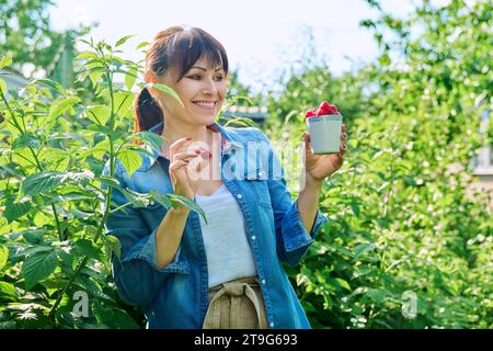 Femme heureuse dans le jardin des framboisiers, avec une tasse de framboises mûres Banque D'Images