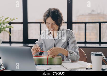Femme joyeuse enveloppant un cadeau de Noël à son bureau avec un ordinateur portable à proximité, sentant l'esprit de vacances Banque D'Images