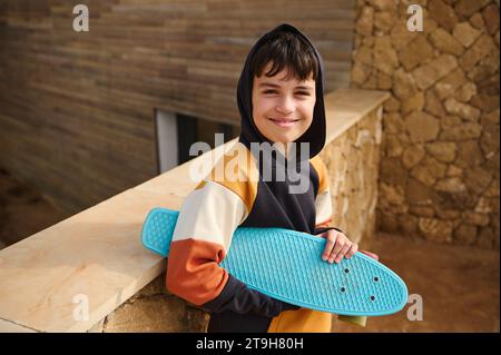 Portrait de style de vie d'un adolescent, hipster, skateboarder dans un sweat à capuche tendance, tenant son skateboard bleu, souriant regardant la caméra, debout à l'extérieur Banque D'Images