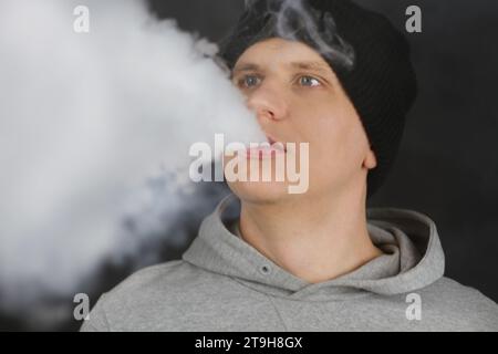 L'homme fume une cigarette électronique sur le fond sombre, les hommes en vapotage à capuche et libère un nuage de vapeur. Guy avec vapotage sur fond noir Banque D'Images