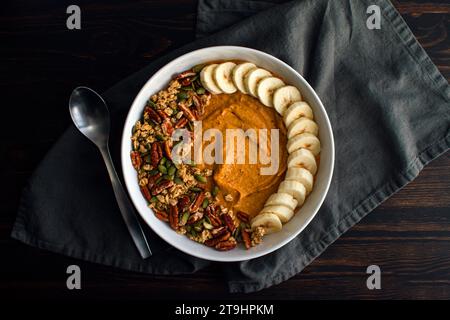 Bol de smoothie à la citrouille sur une table en bois avec une cuillère : smoothie à la citrouille garni de granola, de pacanes, de graines de citrouille et de bananes tranchées Banque D'Images