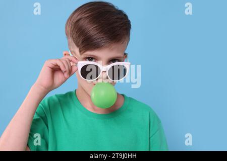 Garçon dans les lunettes de soleil soufflant de la gomme à bulles sur fond bleu clair, espace pour le texte Banque D'Images