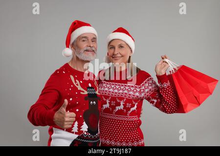 Couple senior en pulls de Noël et chapeaux de Père Noël avec des sacs à provisions sur fond gris Banque D'Images