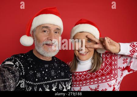 Couple senior en pulls de Noël et chapeaux de Santa prenant selfie sur fond rouge Banque D'Images