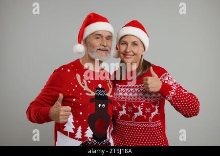 Couple senior dans des pulls de Noël et des chapeaux de Père Noël montrant les pouces vers le haut sur fond gris Banque D'Images