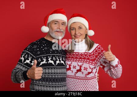 Couple senior dans des pulls de Noël et des chapeaux de Père Noël montrant les pouces vers le haut sur fond rouge Banque D'Images