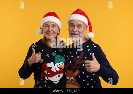 Heureux couple senior dans des pulls de Noël et des chapeaux de Père Noël montrant les pouces vers le haut sur fond orange Banque D'Images