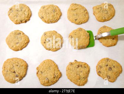 Rangées de biscuits aux pépites de chocolat aux flocons d'avoine fraîchement cuits sur du papier parchemin, spatule en silicone verte soulevant un biscuit de la plaque de cuisson. Banque D'Images