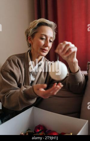 Plan vertical d'une femme d'âge moyen tenant et regardant une boule de Noël festive avec une expression heureuse. La femelle élégante décore l'arbre de Noël pour le nouvel an Banque D'Images