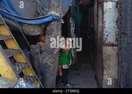 Une jeune fille indienne dans le bidonville de Darukhana, Mazagaon, Mumbai, Inde, souriant à l'étranger de son refuge de bidonville, sa mère regardant prudemment Banque D'Images