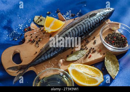 Poisson maquereau cru sur planche à découper sur nappe satinée bleue avec des ingrédients pour la cuisson, citron, épicé, poivrons, sel, huile d'olive. Banque D'Images