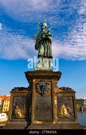 Une statue d'un saint tenant une croix et un livre sur un piédestal. Statue de Jean de Nepomuk sur le pont Charles à Prague contre le ciel. Banque D'Images