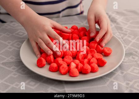 Une assiette ornée de bonbons aux fruits rouges vibrants, saisie avec impatience par les mains d'un enfant, capturant l'essence d'une douceur sucrée Banque D'Images