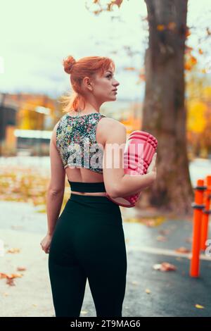 Jeune rousse Caucasian sportswoman tenant un rouleau de mousse dans le parc à la vue arrière d'automne Banque D'Images