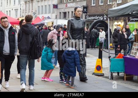 Colchester, Royaume-Uni. 26 novembre 2023. Colchester High Street est fermée à la circulation aujourd'hui pour un marché d'hiver avec des divertissements. Les enfants regardent la statue de Walking Woman. Crédit:Eastern Views/Alamy Live News Banque D'Images