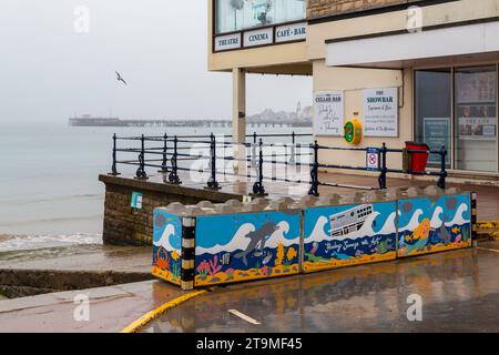 Swanage, Dorset Royaume-Uni. 26 novembre 2023. Météo Royaume-Uni : temps humide, gris et sombre à Swanage dans le Dorset. Peintures sur la barrière temporaire des vagues hivernales de Swanage les blocs de béton mis en place par l’Agence de l’environnement pour réduire l’impact des inondations, ajoutent de la couleur à une journée grise! Crédit : Carolyn Jenkins/Alamy Live News Banque D'Images