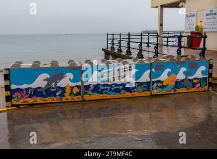 Swanage, Dorset Royaume-Uni. 26 novembre 2023. Météo Royaume-Uni : temps humide, gris et sombre à Swanage dans le Dorset. Peintures sur la barrière temporaire des vagues hivernales de Swanage les blocs de béton mis en place par l’Agence de l’environnement pour réduire l’impact des inondations, ajoutent de la couleur à une journée grise! Crédit : Carolyn Jenkins/Alamy Live News Banque D'Images