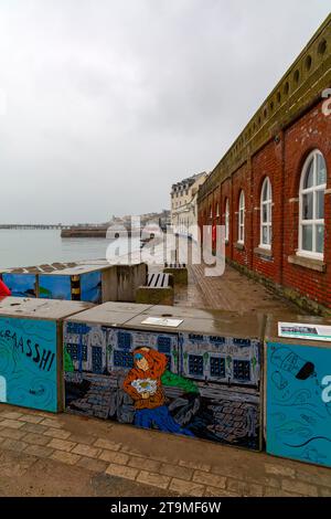 Swanage, Dorset Royaume-Uni. 26 novembre 2023. Météo Royaume-Uni : temps humide, gris et sombre à Swanage dans le Dorset. Peintures sur la barrière temporaire des vagues hivernales de Swanage les blocs de béton mis en place par l’Agence de l’environnement pour réduire l’impact des inondations, ajoutent de la couleur à une journée grise! Crédit : Carolyn Jenkins/Alamy Live News Banque D'Images