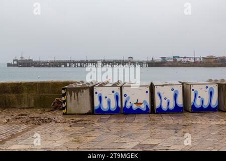 Swanage, Dorset Royaume-Uni. 26 novembre 2023. Météo Royaume-Uni : temps humide, gris et sombre à Swanage dans le Dorset. Peintures sur la barrière temporaire des vagues hivernales de Swanage les blocs de béton mis en place par l’Agence de l’environnement pour réduire l’impact des inondations, ajoutent de la couleur à une journée grise! Crédit : Carolyn Jenkins/Alamy Live News Banque D'Images