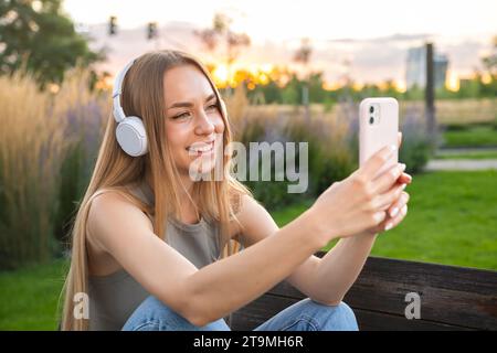 Le blogueur blonde tourne des histoires assis dans le parc sur le banc. Une jeune femme au casque prend un selfie. Banque D'Images