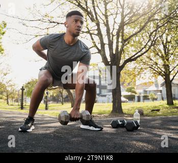 Adaptez jeune homme noir dans des vêtements de sport et des écouteurs sans fil faisant des squats avec des haltères dans le parc Banque D'Images