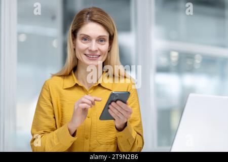 Portrait rapproché d'une jeune femme, employée de bureau, femme d'affaires assise dans le bureau à la table et utilisant le téléphone, souriant à la caméra. Banque D'Images